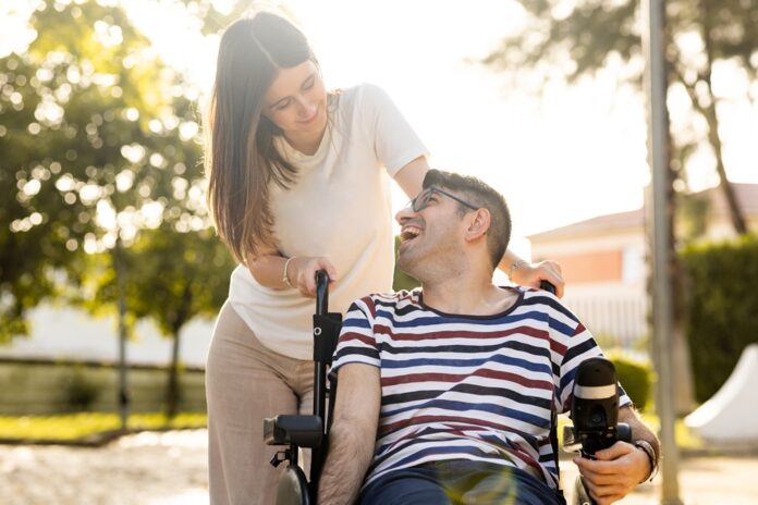 An adult man with a disability sits in an automatic wheelchair with a young woman in a park at sunset.The woman and the man look at each other happily. Concept of support for disabled people. Disability